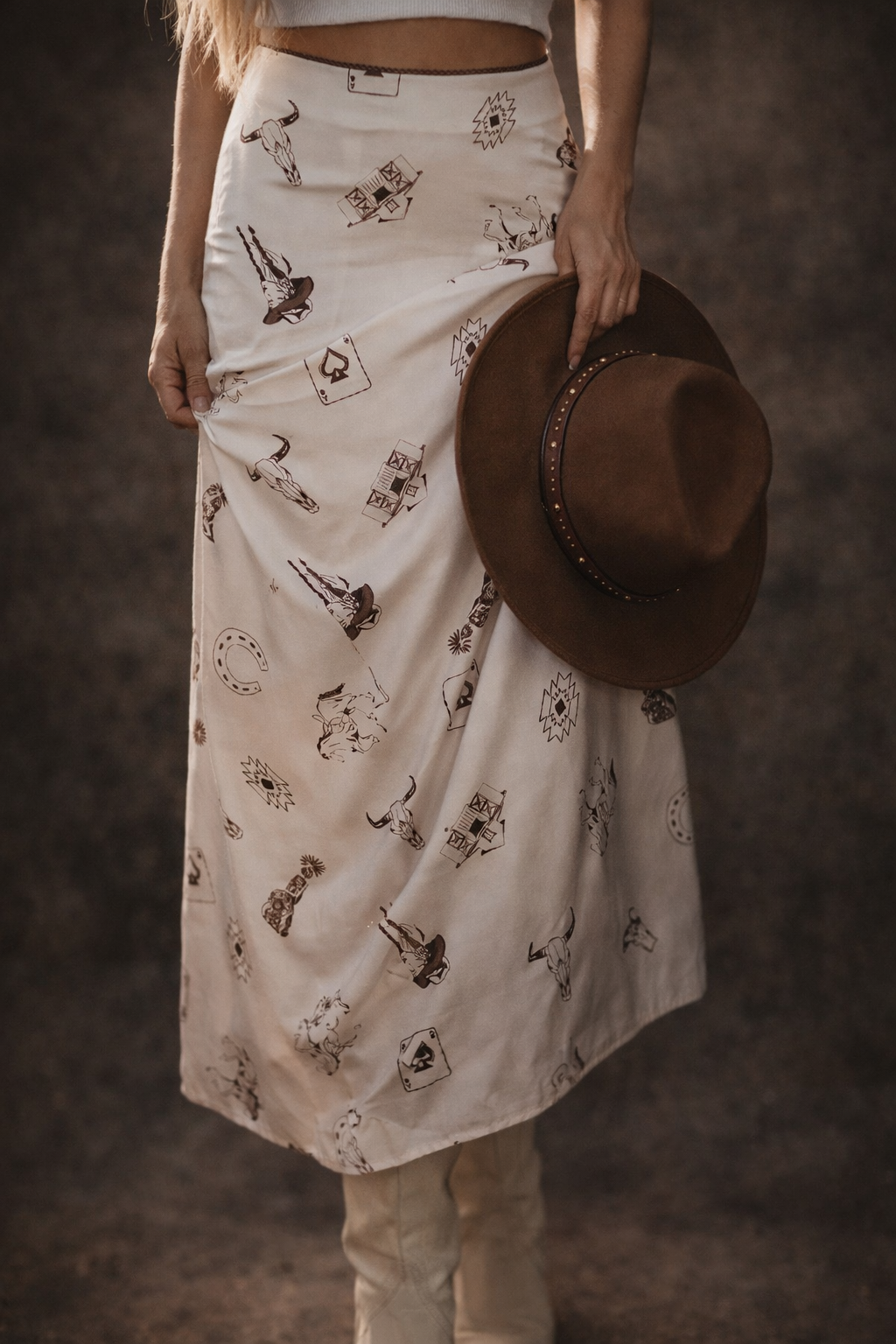 Woman wearing a western print midi skirt with cowgirl-inspired graphics, cowboy boots, and a brown western hat for Westynwild boutique.